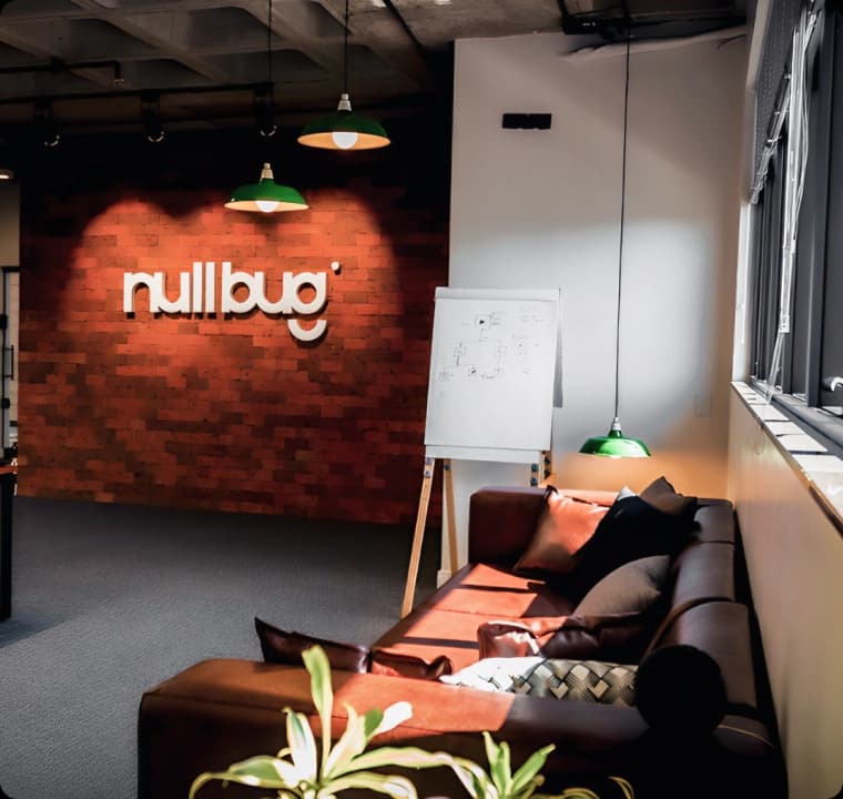 Nullbug Software Development headquarters. In the photo you can see a brown sofa with cushions and in front you can see the white Nullbug writing in high relief on a wall made of industrial bricks.
