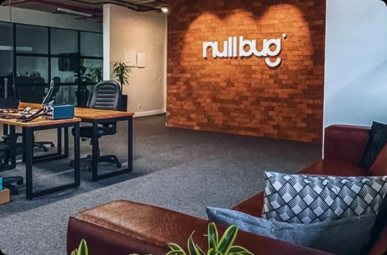 Nullbug headquarters. In the photo you can see a brown sofa with cushions and in front you can see the word Nullbug in relief on a wall made of industrial bricks. In front we also have industrial-style work desks for software developers and specialized programmers.